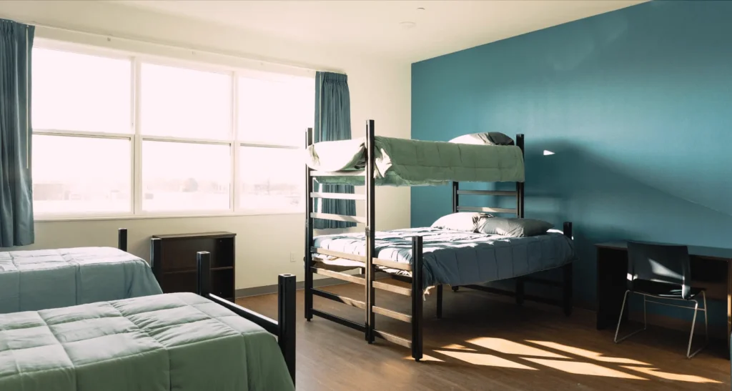 A bright dorm room with two sets of bunk beds, each with blue-green bedding, and two desks with chairs. Sunlight streams in through a large window, casting shadows on the wood floor.
