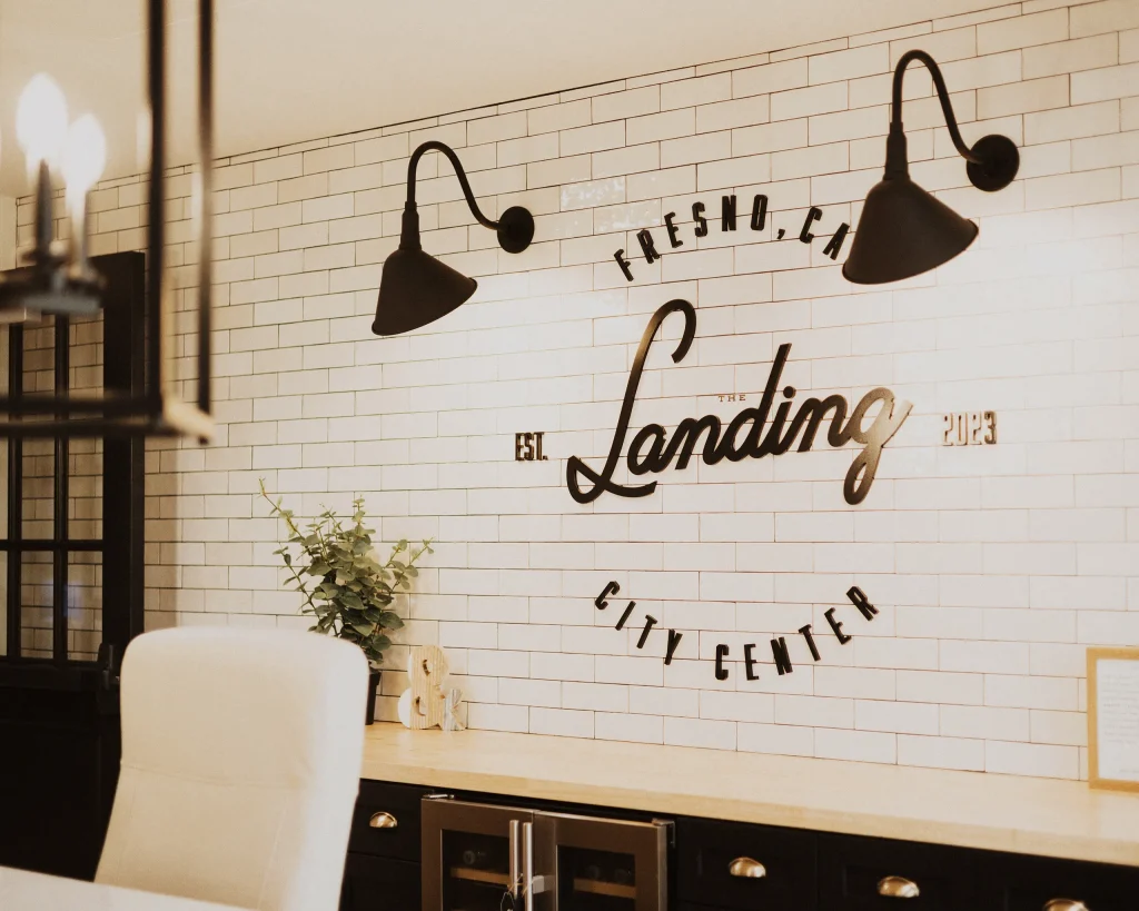 A modern office space with a white brick wall featuring black script reading The Landing, Fresno, CA, City Center, Est. 2023. Black wall lamps and a white chair are visible, with plants and decor on the desk.