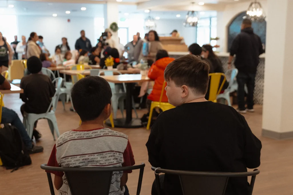 Two boys sit side by side on chairs, facing a busy room filled with people of various ages who are gathered around tables, socializing in a bright, modern space.