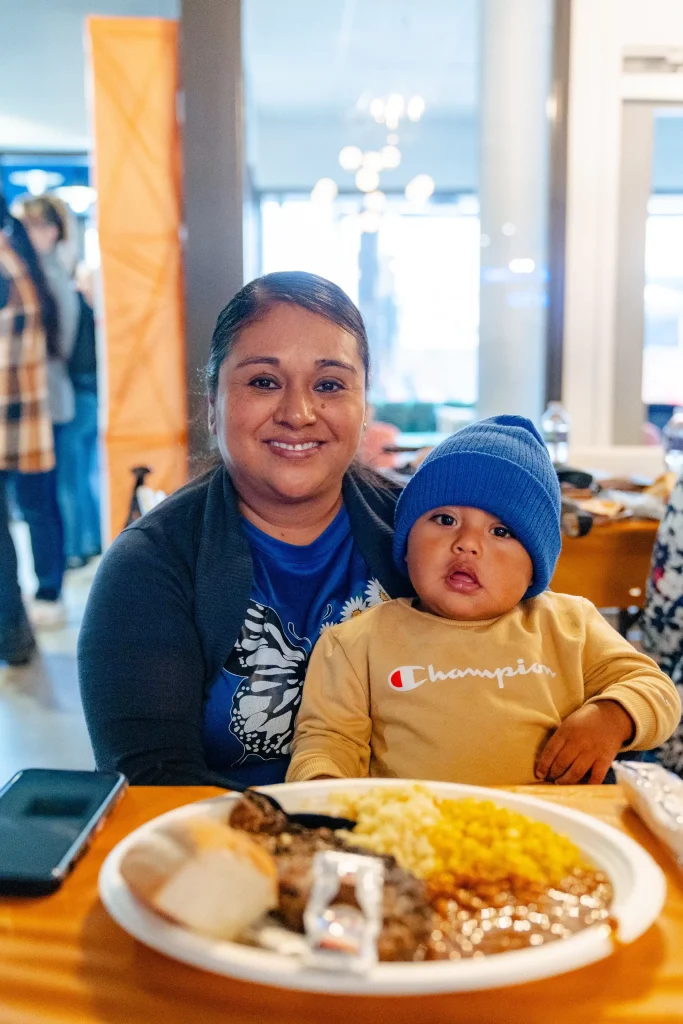 A woman smiling and holding a young child in a blue beanie, seated at a table with a plate of food including bread, corn, and meat. The setting appears to be a casual indoor gathering.