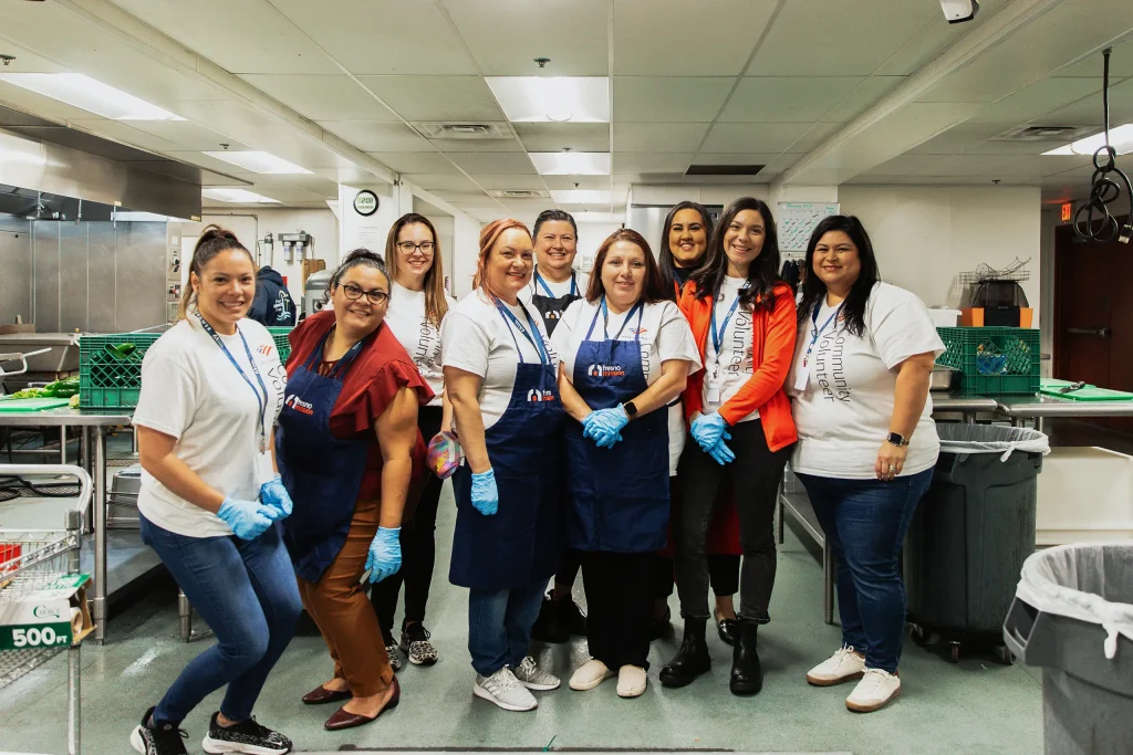A group of eleven people, some in aprons and gloves, smile together in a commercial kitchen, suggesting they are part of a volunteer or community service activity.