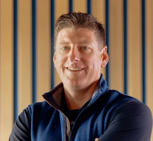 A man with short brown hair wearing a dark blue zip-up vest and black shirt stands smiling in front of a background with vertical wooden slats.