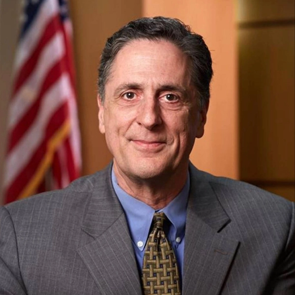 A middle-aged man in a gray suit, blue shirt, and patterned tie smiles at the camera. An out-of-focus American flag is visible in the background.