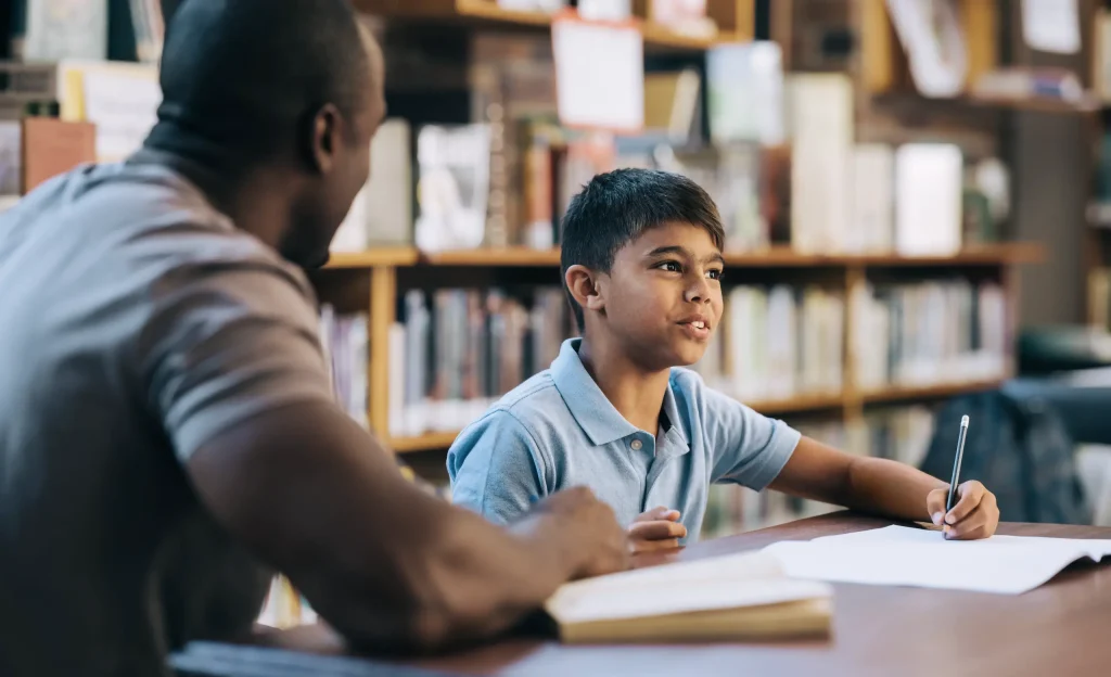 A young boy sits at a table in a library, holding a pencil and talking to an adult man beside him. Bookshelves filled with books are visible in the background.