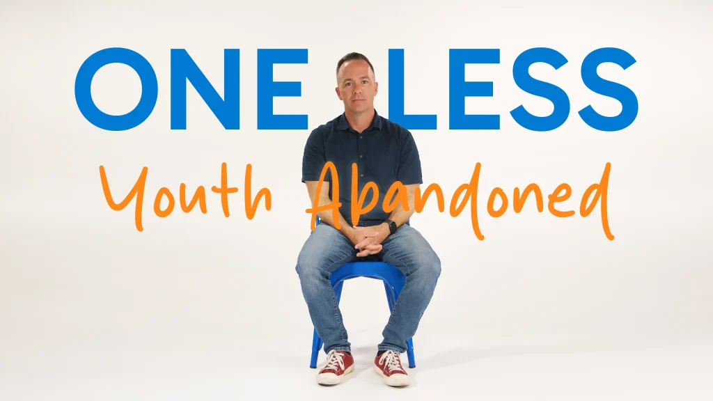A man sits on a blue stool against a white background. Large blue text above him reads ONE LESS and orange text in front reads Youth Abandoned. He wears a dark shirt, jeans, and red sneakers.