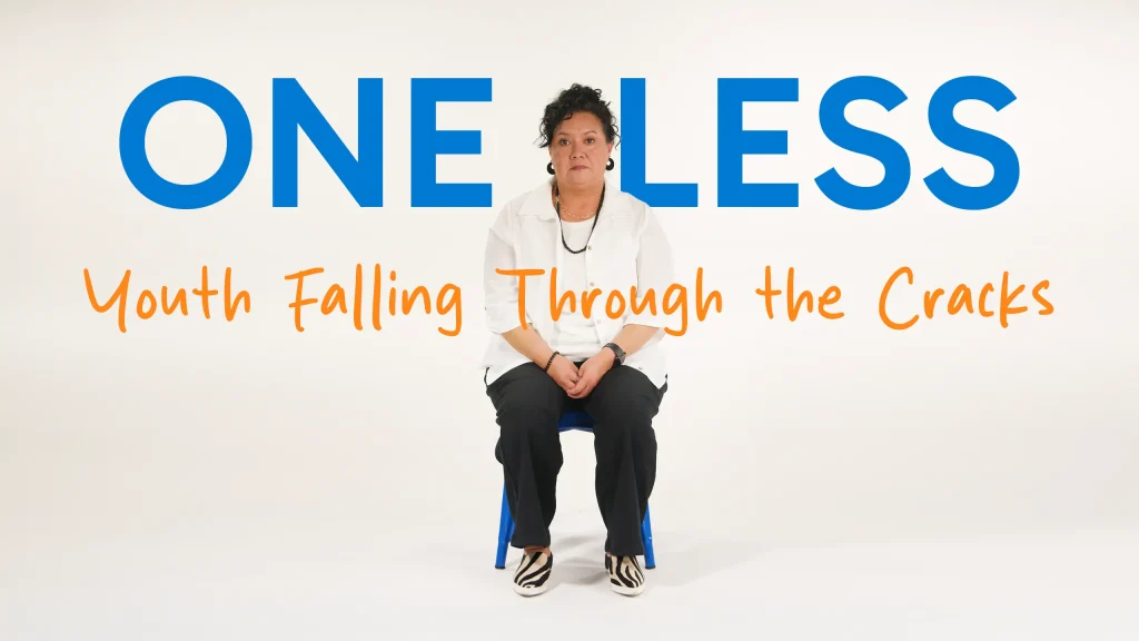 A person sits on a blue chair against a white background. Large blue text behind reads “ONE LESS,” with orange text below saying “Youth Falling Through the Cracks.”.