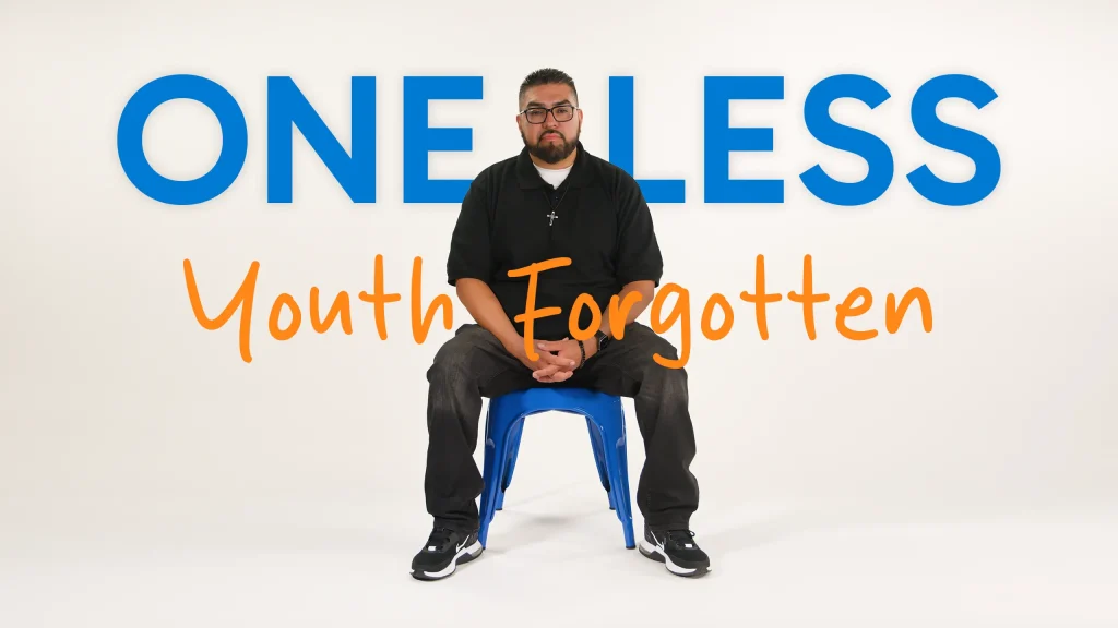 A man sits on a blue chair against a white background with the text ONE LESS Youth Forgotten in large blue and orange letters behind him.