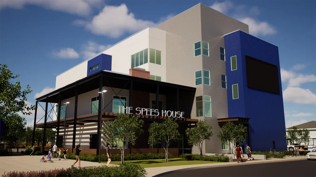 A modern multi-story building with a blue and gray exterior, large windows, and a sign reading THE SPEES HOUSE. People are walking on the sidewalk and there are trees and plants in front. The sky is partly cloudy.