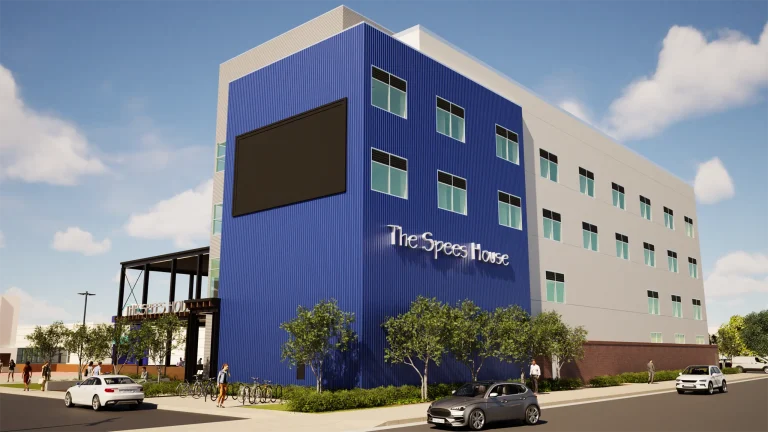 A modern, three-story building with blue and gray panels, large windows, and a sign reading The Spees House. People walk and bike nearby, and cars are parked on the street under a sunny sky with scattered clouds.
