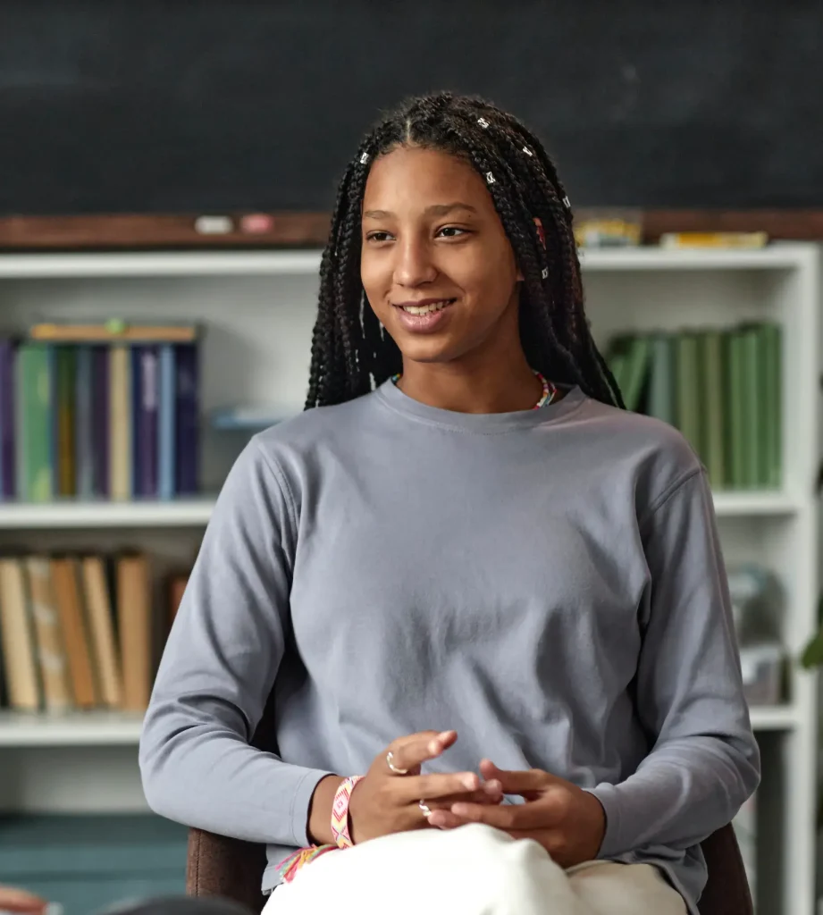 A young woman with braids sits and smiles during a conversation with another person in a room with shelves of colorful books in the background.